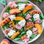 The Grilled Peach and Burrata Salad features succulent grilled peach slices, creamy burrata cheese, and savory prosciutto on a bed of fresh greens. With charred grill marks on the peaches and a sprinkle of cracked black pepper, this dish rests elegantly on a wooden table.