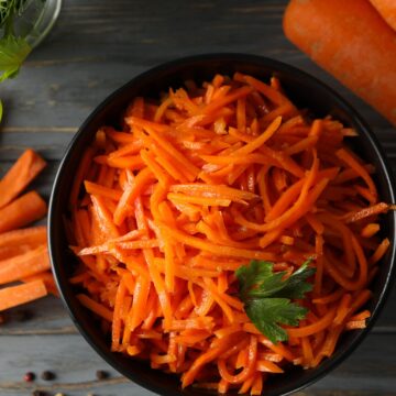 Shredded carrots in a black bowl with a sprig of parsley. There is also a couple of carrots off to the side.