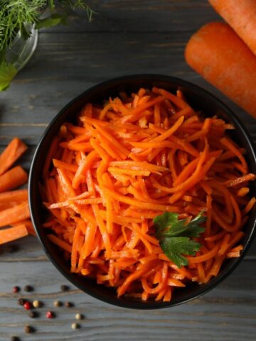 Shredded carrots in a black bowl with a sprig of parsley. There is also a couple of carrots off to the side.