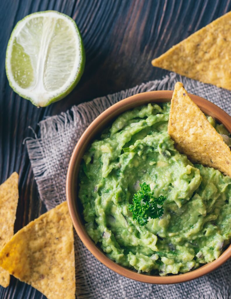 Guacamole in a brown bowl with tortilla chips next to it and a lime on a brown table cloth.