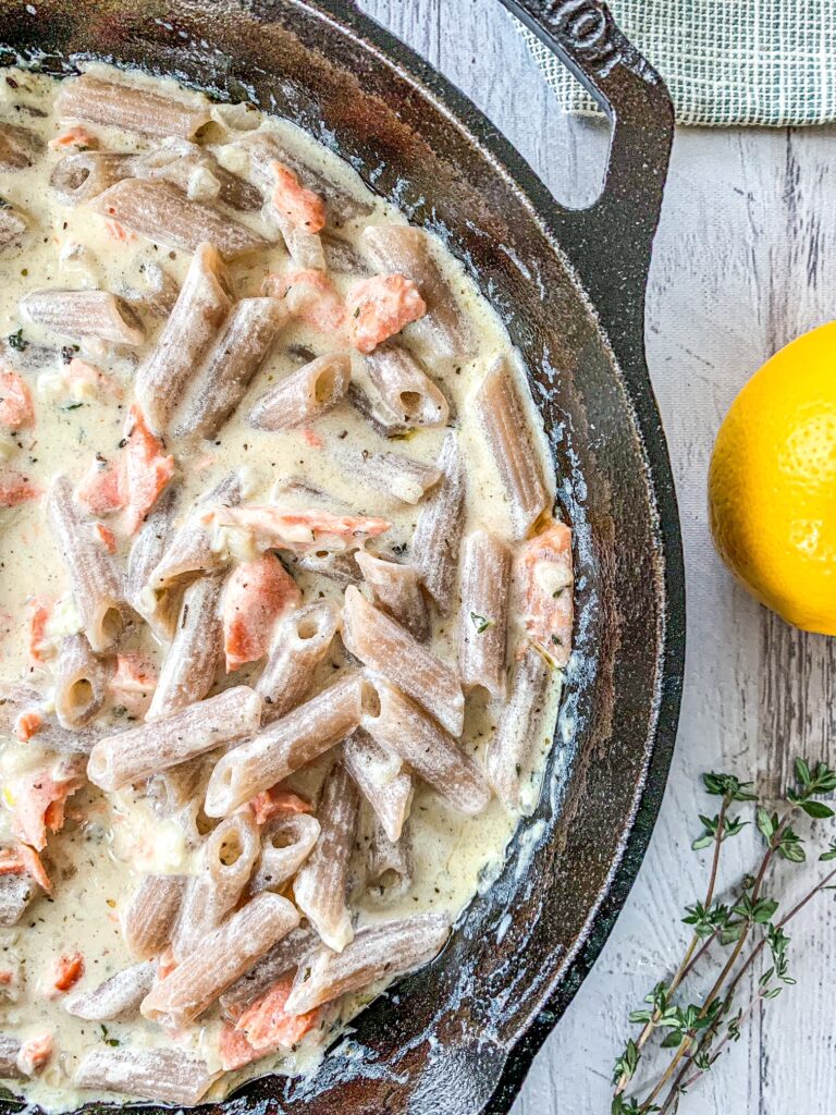 Penne pasta and salmon in a cream sauce in a cast iron skillet next to lemon and thyme.