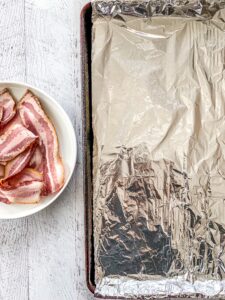 A baking sheet lined with tin foil and uncooked bacon on a white dish.