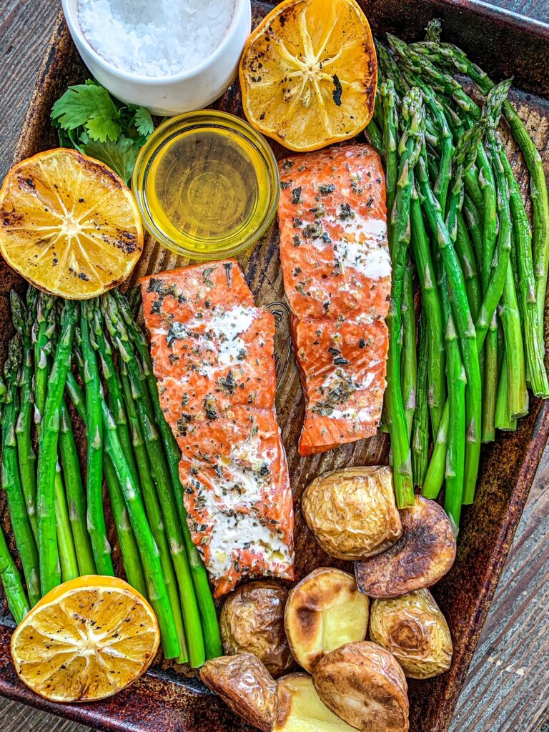 Two pieces of grilled salmon on a baking sheet surrounded with asparagus, potatoes and grilled lemons.