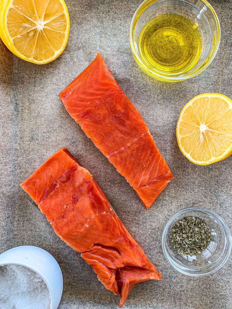 Wild uncooked salmon on parchment paper with oil, seasonings, lemon and salt in small bowls.