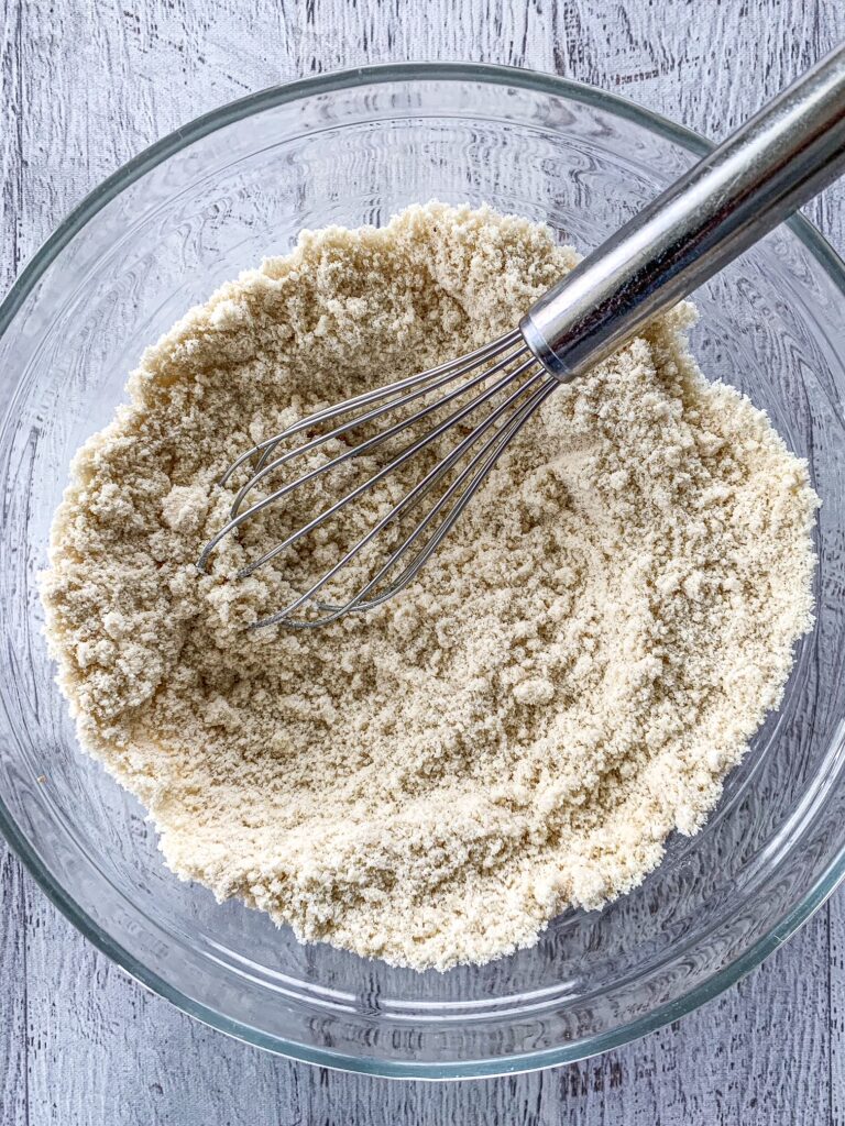 Almond flour, gluten-free flour and baking soda combined in a glass bowl with a silver whisk.