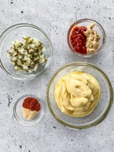 A bowl of mayonnaise, a bowl of paprika and garlic powder, a bowl of ketchup and mustard and a bowl of pickles on a white backdrop.