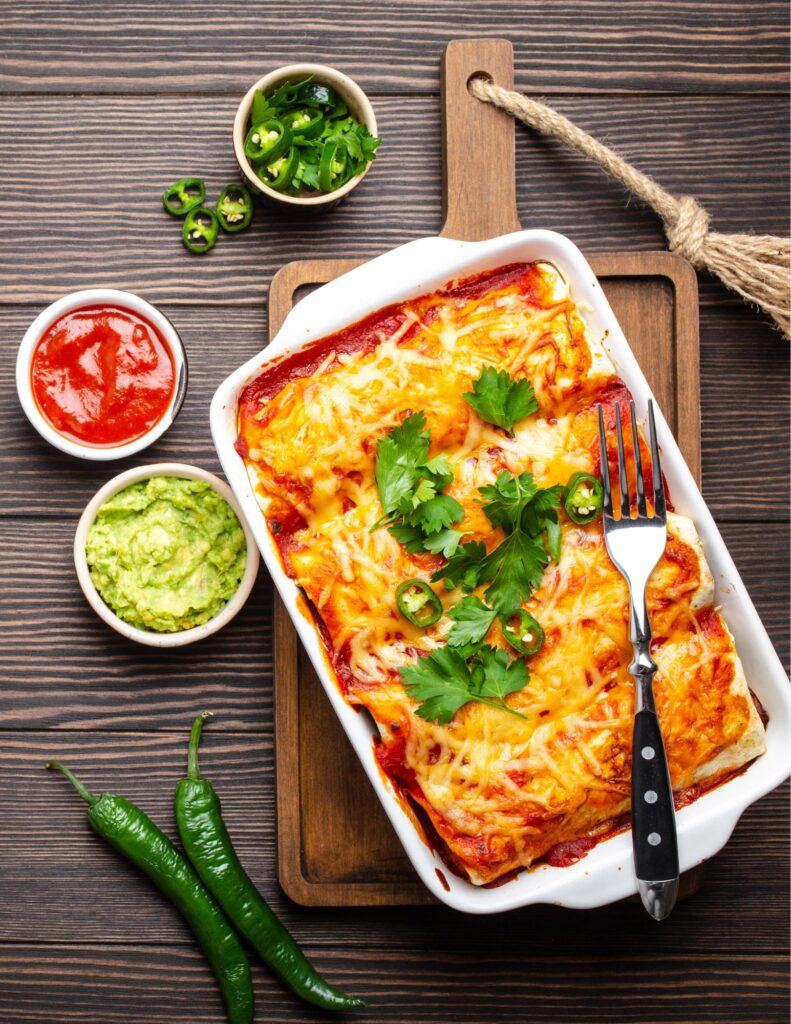 Enchiladas in red sauce topped with orange cheddar cheese and cilantro. Two small bowls are next to the white baking dish, one has guacamole and one has salsa.