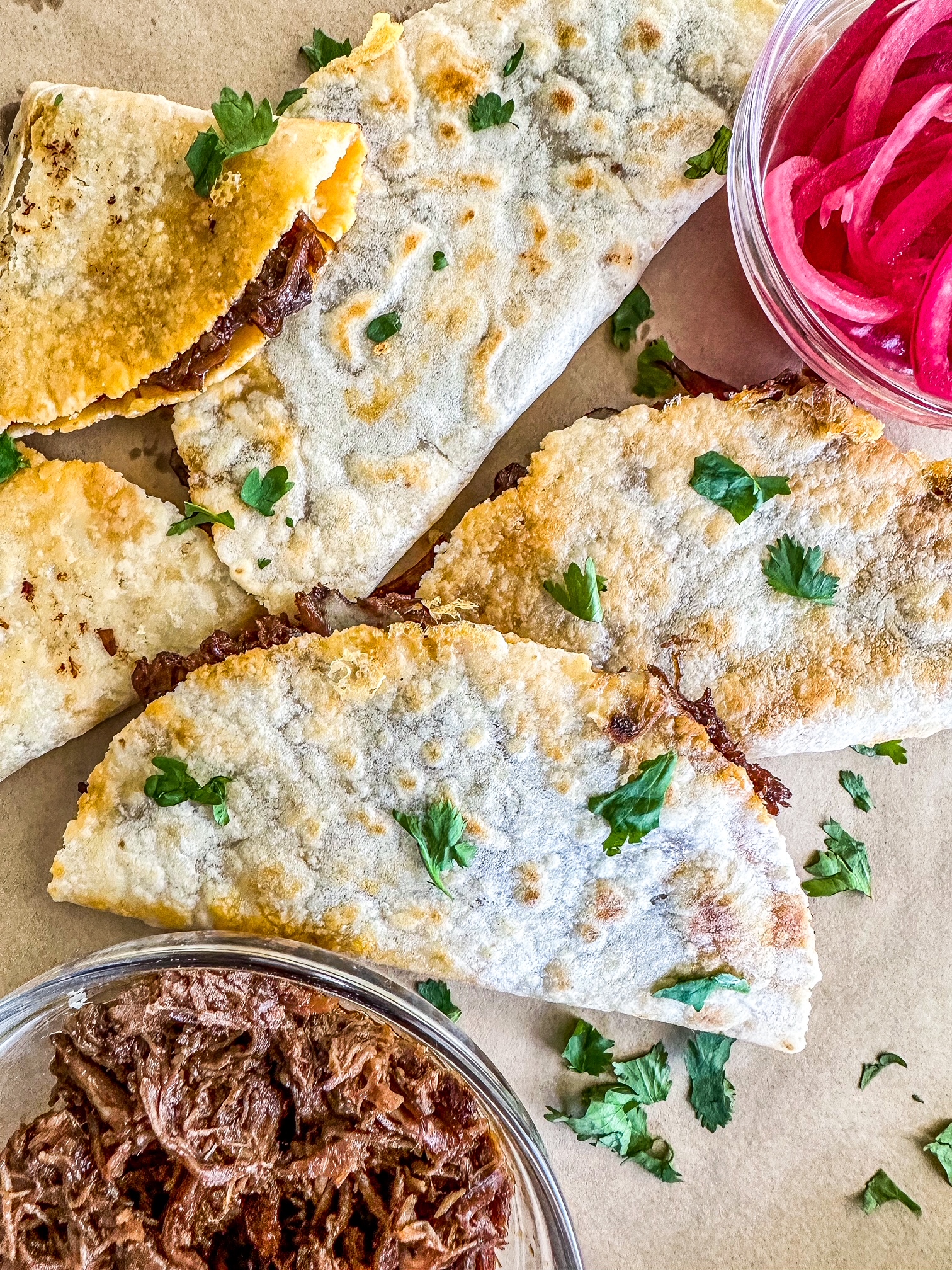 Barbacoa Quesadillas on a sheet of parchment paper with shredded barbacoa in a bowl