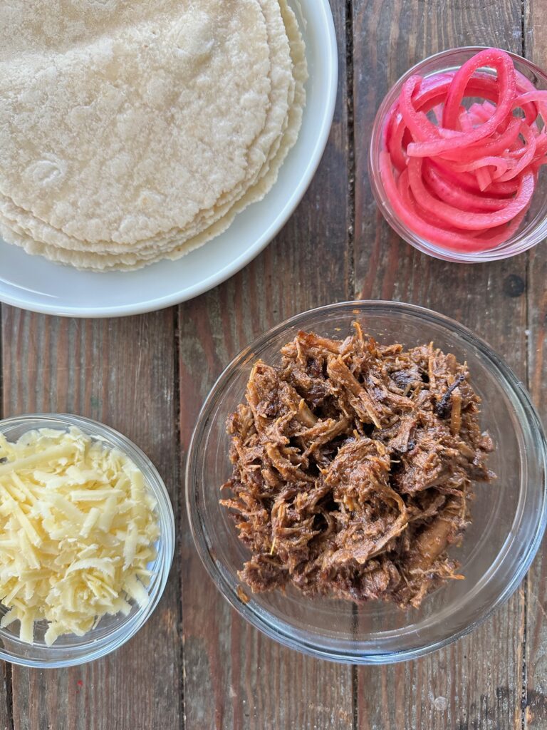 Barbacoa Quesadilla ingredients. 1 bowl of shredded barbacoa, 1 bowl of shredded white cheddar cheese, tortillas and a bowl of picked red onions on a brown background.