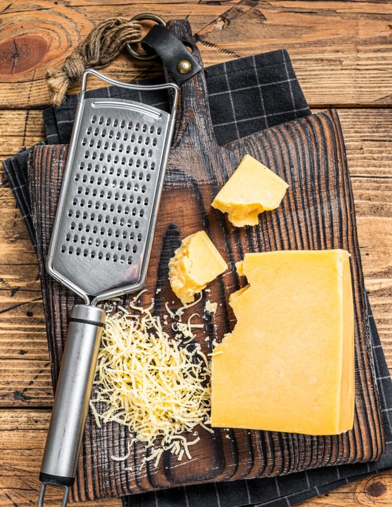 White cheddar cheese grated on a wood cutting board with a hand held grater and a block of cheese.