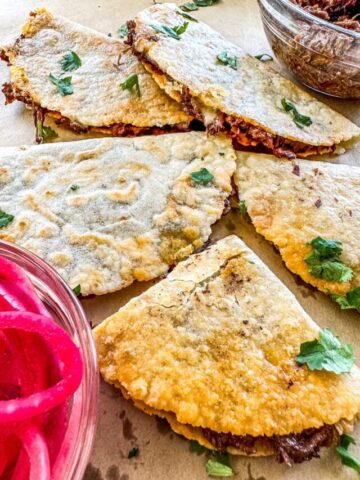 Barbacoa Quesadillas on a sheet of parchment paper. Pink pickled onions and shredded barbacoa in the background.