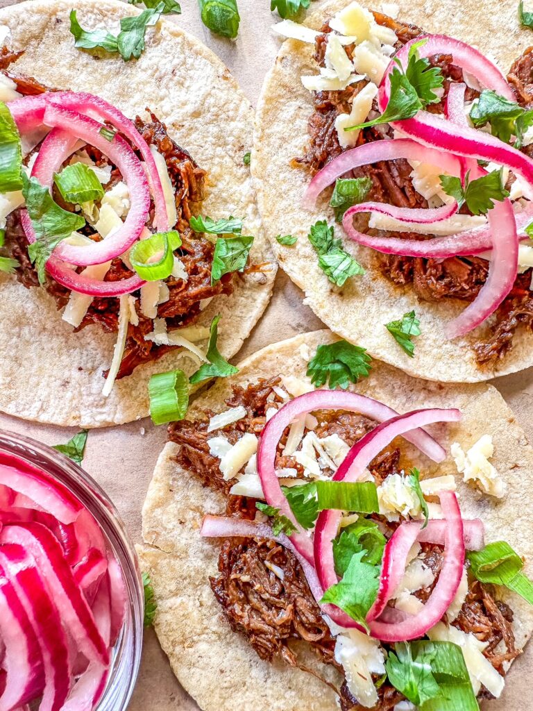3 Barbacoa tacos on corn tortillas with picked red onions and cilantro on top.