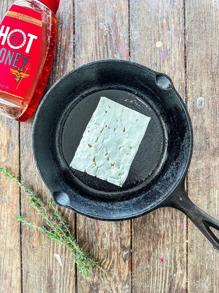 Block of feta cheese in a cast iron skillet on a wood background. Hot honey and thyme are next to the skillet.