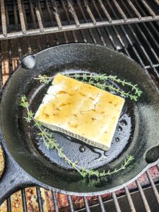 Block of feta cheese in a cast iron skillet with honey and thyme leaves on the grill grates of a pellet smoker.