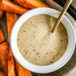 A bowl of creamy hot honey mustard sauce with a golden spoon in it, surrounded by cooked sweet potato wedges on a dark tray.