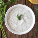 Lemon yogurt dill sauce in a white bowl with dill and lemon next to it on a wood table.