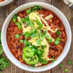 Overhead shot of dutch oven chili topped with white cheddar cheese, green onions cilantro and jalepeno.