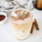 Iced maple latte in a glass with a glass straw. Two cinnamon sticks in the background.