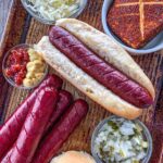 Smoked hot dogs in a bun with toppings surrounding it.