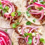 Tacos de Barbacoa on parchment paper. Topped with white cheddar and pink pickled onions.