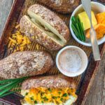 Traeger baked potatoes on a sheet pan. One potato is cut open and filled with cheese and chives.