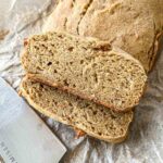 Close-up of sliced homemade bread on parchment paper. The loaf is golden brown with a dense, textured interior, revealing visible specks of grains or seeds. A knife handle is partially visible next to the bread.