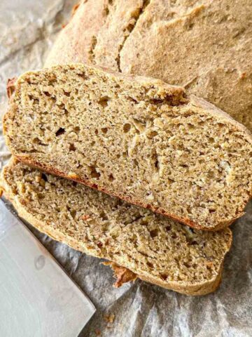 Close-up of sliced homemade bread on parchment paper. The loaf is golden brown with a dense, textured interior, revealing visible specks of grains or seeds. A knife handle is partially visible next to the bread.