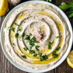 A creamy bowl of hummus drizzled with olive oil, sprinkled with spices, and garnished with fresh parsley. Lemon slices and cloves of garlic are partially visible around the bowl, set on a wooden table.