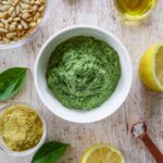 A bowl of green pesto is surrounded by pine nuts, olive oil, basil leaves, a half lemon, nutritional yeast, and a small copper spoon with salt, all on a wooden surface.