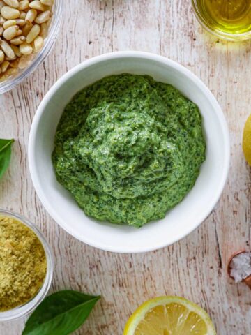 A bowl of green pesto is surrounded by pine nuts, olive oil, basil leaves, a half lemon, nutritional yeast, and a small copper spoon with salt, all on a wooden surface.