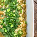 A dish of enchiladas topped with grated cheese, sliced jalapeños, and fresh cilantro in a white baking dish. The enchiladas are covered in green sauce and garnished with chopped green onions.
