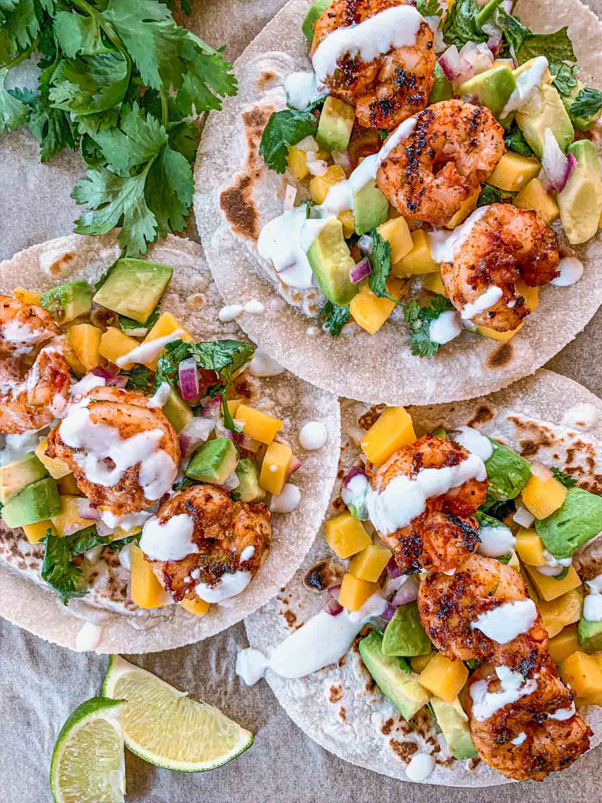 Three shrimp tacos on tortillas topped with diced mango, avocado, red onion, cilantro, and a drizzle of creamy white sauce. A lime wedge and cilantro garnish are nearby.