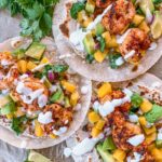 Three grilled shrimp tacos topped with diced mango, avocado, red onion, fresh cilantro, and drizzled with a creamy white sauce. Lime wedges and cilantro garnish the presentation on a rustic surface.