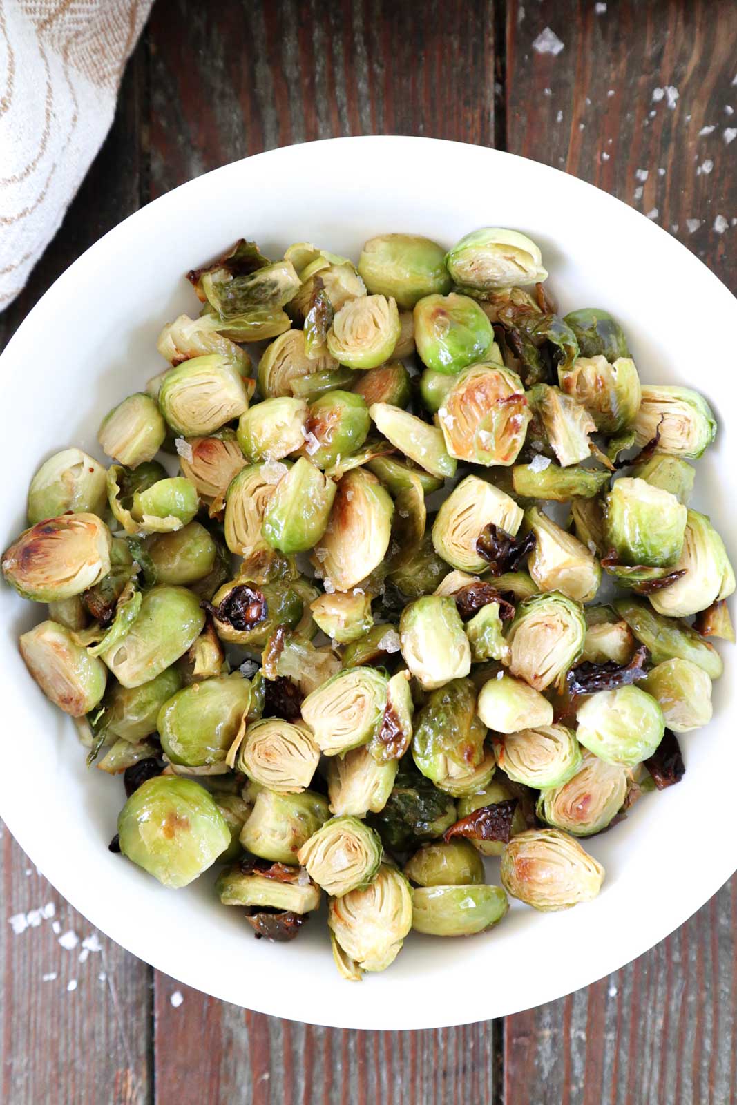 A bowl of roasted Brussels sprouts seasoned with coarse salt sits on a wooden surface. The sprouts are halved, showing a golden-brown and crispy appearance.