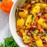 A close-up of a tropical fruit salad in a white bowl featuring fresh mango chunks, diced red onions, and chopped cilantro mixed with small orange pepper pieces. An orange pepper and cilantro leaves are on the white wooden surface nearby.