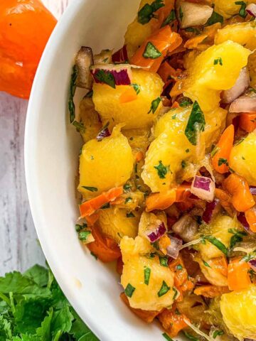 A close-up of a tropical fruit salad in a white bowl featuring fresh mango chunks, diced red onions, and chopped cilantro mixed with small orange pepper pieces. An orange pepper and cilantro leaves are on the white wooden surface nearby.