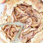 A rustic pear galette with a golden, flaky crust and spiced pear slices arranged in the center. A wedge is cut out, revealing the tender fruit and crumbly edges, on a parchment-paper-lined surface.