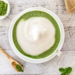 A creamy matcha latte in a white cup on a white wooden surface, with frothy milk swirling through the vibrant green beverage. A wooden spoon with matcha powder and a small dish of matcha are beside it, along with a bamboo whisk and mint leaves.