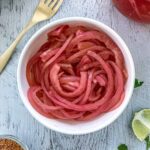 A white bowl filled with pickled red onions sits on a light-colored wooden table. A gold fork is placed nearby, and the edge of a lime and a small dish of spices are visible.