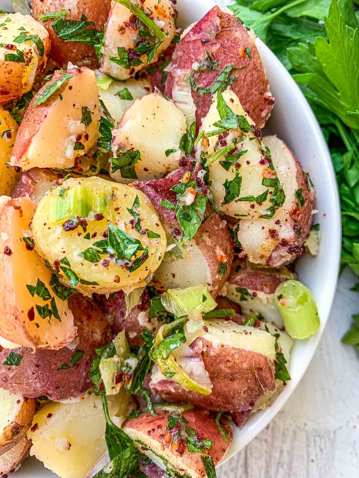 A close-up of a potato salad in a white bowl. The salad features chunks of red and yellow potatoes, chopped green onions, parsley, and a seasoning of spices. Fresh parsley is visible in the background, enhancing the dishs fresh appearance.