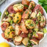 A close-up of a bowl filled with a colorful potato salad. The salad consists of red and yellow potato chunks mixed with chopped herbs, celery pieces, and seasoning. Lemon and parsley are visible in the background.