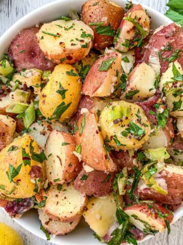 A close-up of a bowl filled with a colorful potato salad. The salad consists of red and yellow potato chunks mixed with chopped herbs, celery pieces, and seasoning. Lemon and parsley are visible in the background.