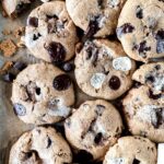A batch of freshly baked s'mores chocolate chip cookies with chunks of chocolate and visible marshmallow bits, spread across a parchment-lined surface. The cookies are golden brown with a slightly crispy texture.