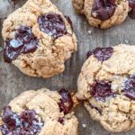Close-up of three freshly baked chocolate chip cookies on a textured surface. The cookies are thick, with visible chunks of melted chocolate and a sprinkle of coarse sea salt on top.