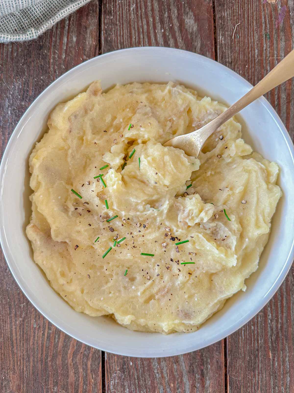 A bowl of creamy mashed potatoes garnished with chopped chives and freshly cracked black pepper. A spoon is resting in the dish, which is set on a rustic wooden table.