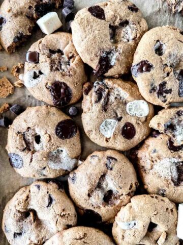 A batch of freshly baked cookies with chunks of chocolate and marshmallows on a parchment-lined surface. The cookies are golden brown and feature a variety of textures from the melted chocolate and marshmallow pieces.