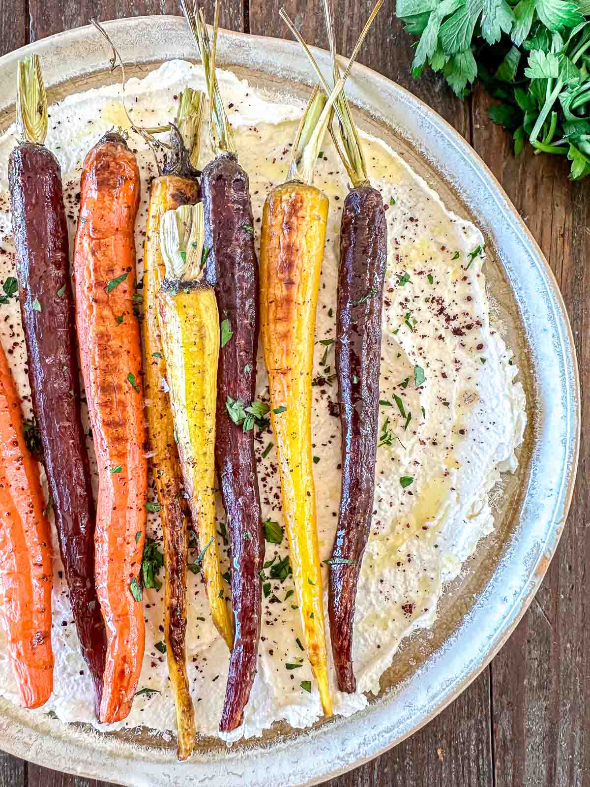 A platter features roasted rainbow carrots, including orange, purple, and yellow varieties, laid on a bed of creamy hummus. The dish is garnished with fresh herbs, all served on a rustic wooden surface.