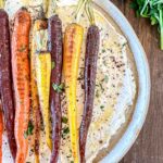 Colorful roasted carrots on a round white plate with a whipped feta with herbs. A bunch of fresh parsley is visible in the top right corner, all set on a wooden table.