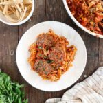 A white plate filled with tagliatelle pasta topped with three meatballs and tomato sauce sits on a wooden table. Fresh parsley is nearby, with uncooked pasta and a striped cloth in the background.