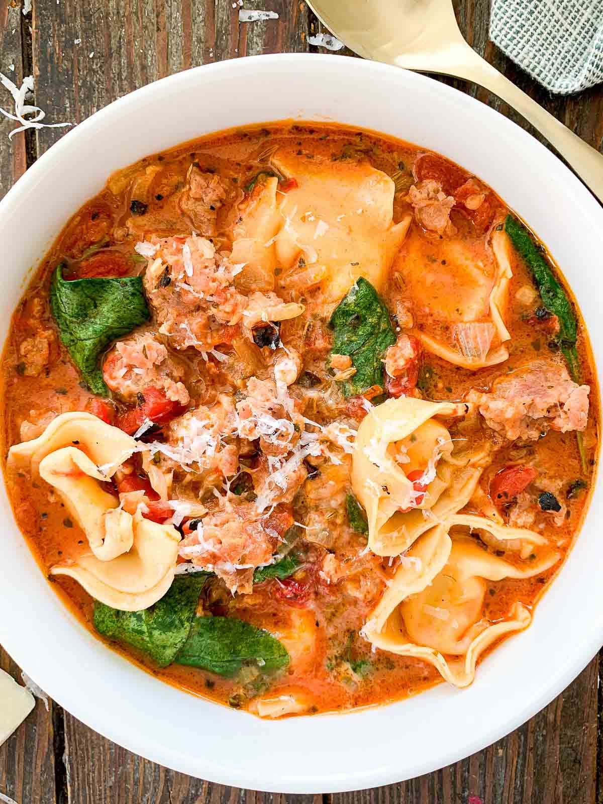 A bowl of creamy tomato tortellini soup with chunks of sausage, spinach, and topped with grated cheese. The soup has a rich, savory appearance and is served in a white bowl placed on a wooden table. A gold spoon is partially visible beside the bowl.